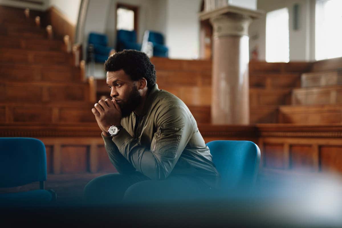 A single man sits in amongst empty chairs and thinks before a group therapy session